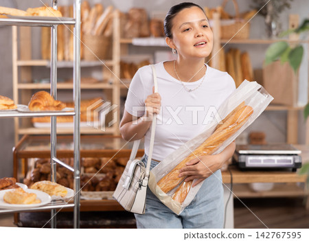 Asian woman buyer with baguettes in plastic package stands in trading floor of bakery Asian woman buyer with baguettes in plastic package stands in trading floor of bakery 142767595