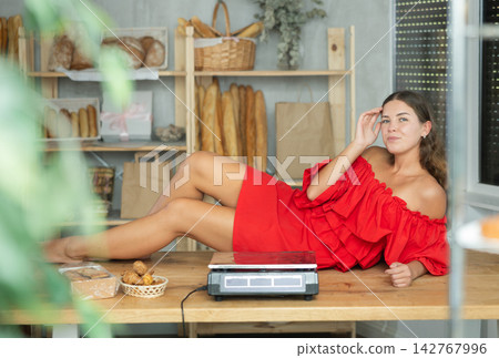 Girl in red dress with bare shoulders is lying on table in trading floor of bakery. Girl in red dress with bare shoulders is lying on table in trading floor of bakery. 142767996