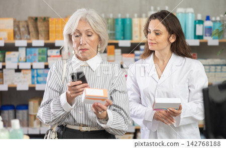 Senior woman in pharmacy scans QR code on omega 3 supplement, woman pharmacist in background Senior woman in pharmacy scans QR code on omega 3 supplement, woman pharmacist in background 142768188