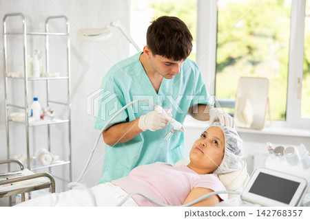 Young male cosmetologist doing moisturizing procedure to young woman 142768357