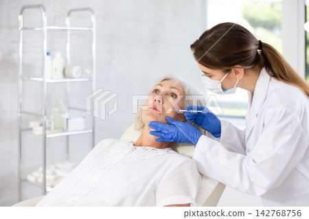 Eldery woman is getting a rejuvenating facial injections. She is lying calmly at clinic. Eldery woman is getting a rejuvenating facial injections. She is lying calmly at clinic. 142768756