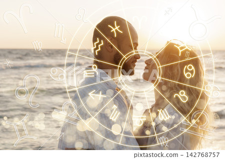 Couple looking at each other with zodiac signs on the background near the sea during sunset Couple looking at each other with zodiac signs on the background near the sea during sunset 142768757