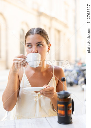 Girl is sitting at table in outdoor cafe, enjoying delicious cup of coffee Girl is sitting at table in outdoor cafe, enjoying delicious cup of coffee 142768827