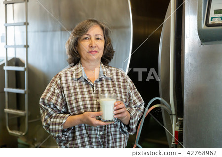 Portrait of a elderly woman standing in the dairy farm, holding a glass of milk Portrait of a elderly woman standing in the dairy farm, holding a glass of milk 142768962