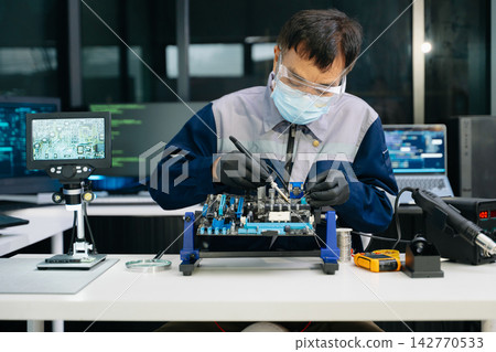 Skilled Asian engineer in safety gear tests a computer motherboard with a multimeter. Precision electronic repair and troubleshooting in a modern manufacturing laboratory. 142770533