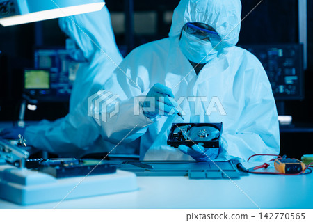 Cleanroom technicians in protective suits repairing a computer hard drive. Engineers conducting digital forensics and data recovery Cleanroom technicians in protective suits repairing a computer hard drive. Engineers conducting digital forensics and data recovery 142770565