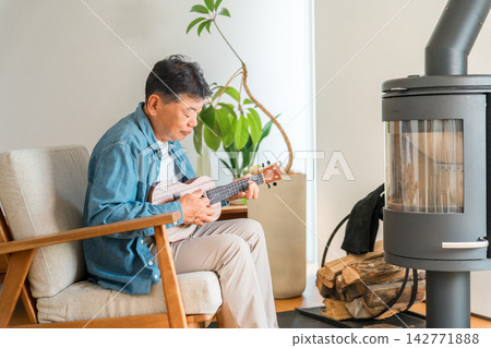 A senior man enjoys playing the ukulele in his home with a wood-burning stove. Enjoying his hobby in winter. A senior man enjoys playing the ukulele in his home with a wood-burning stove. Enjoying his hobby in winter. 142771888