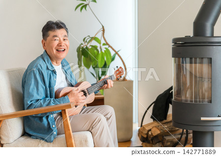 A senior man enjoys playing the ukulele in his home with a wood-burning stove. Enjoying his hobby in winter. A senior man enjoys playing the ukulele in his home with a wood-burning stove. Enjoying his hobby in winter. 142771889