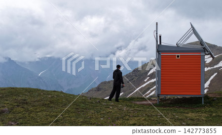 Man in black outfit near orange weather station in mountainous landscape Man in black outfit near orange weather station in mountainous landscape 142773855
