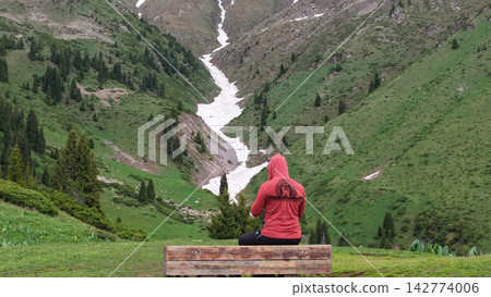 Person in red hoodie sitting on bench overlooking mountainous landscape Person in red hoodie sitting on bench overlooking mountainous landscape 142774006