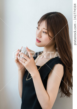 A woman calmly relaxes with a cup in her hand, wearing elegant fashion with a matching necklace and ring. A woman calmly relaxes with a cup in her hand, wearing elegant fashion with a matching necklace and ring. 142774384
