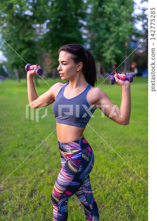 Athletic young woman exercising with purple dumbbells on the grass Athletic young woman exercising with purple dumbbells on the grass 142775285
