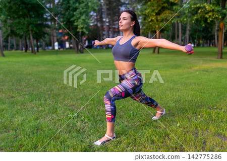 Young woman performing a lunge with dumbbells in a green park Young woman performing a lunge with dumbbells in a green park 142775286