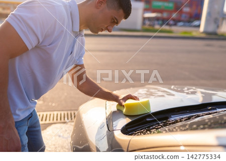 Young man washes his car using a large porous sponge Young man washes his car using a large porous sponge 142775334
