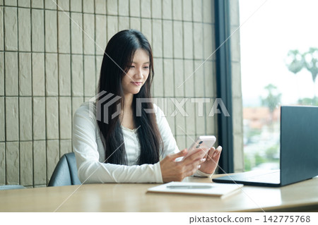Gen Z Young Woman Using Smartphone at Desk with Laptop in Modern Workspace Gen Z Young Woman Using Smartphone at Desk with Laptop in Modern Workspace 142775768