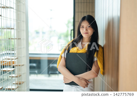 Young Woman Student Holding Black Folder Standing in Modern University Hallway Smiling Confidently 142775769