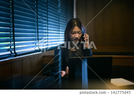 Woman Talking on Phone While Working on Laptop in Dark Moody Cafe Booth Woman Talking on Phone While Working on Laptop in Dark Moody Cafe Booth 142775845
