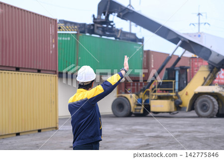 Industrial logistics worker directing container lifting operation at cargo shipping terminal Industrial logistics worker directing container lifting operation at cargo shipping terminal 142775846