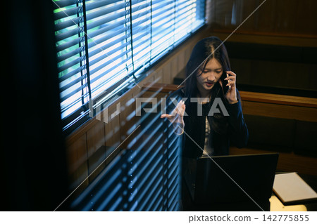 Gen Z Young Woman Talking on Phone While Working on Laptop in Dark Moody Cafe Booth Gen Z Young Woman Talking on Phone While Working on Laptop in Dark Moody Cafe Booth 142775855