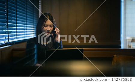 Gen Z Young Woman Talking on Phone While Working on Laptop in Dark Moody Cafe Booth Gen Z Young Woman Talking on Phone While Working on Laptop in Dark Moody Cafe Booth 142775898