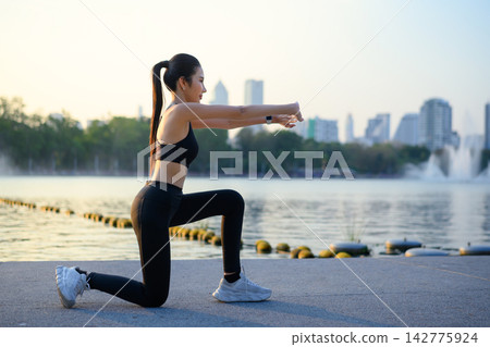 Woman raises arms stretching by lake with city view at sunrise Woman raises arms stretching by lake with city view at sunrise 142775924