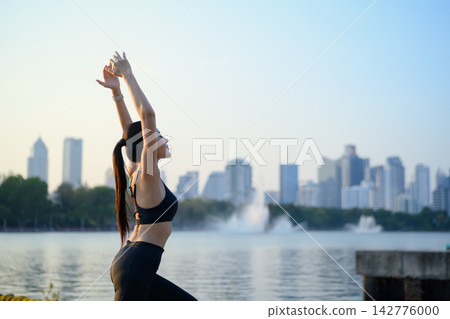 Woman raises arms stretching by lake with city view at sunrise Woman raises arms stretching by lake with city view at sunrise 142776000