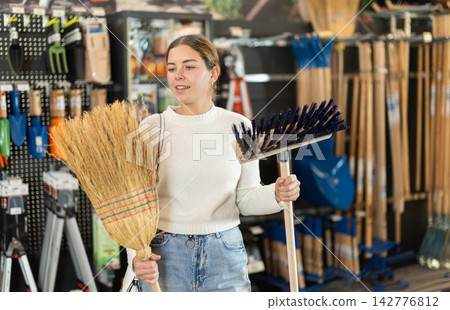 Girl in hardware store examines broom and besom. Girl in hardware store examines broom and besom. 142776812