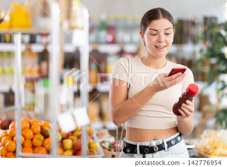 European young woman customer at shop scans QR code on ketchup bottle using phone camera European young woman customer at shop scans QR code on ketchup bottle using phone camera 142776954
