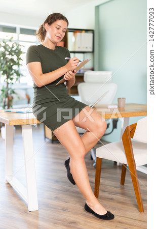 Young woman sits on desk with clipboard Young woman sits on desk with clipboard 142777093