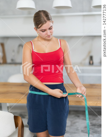 Teenage girl measures her waist and chest with measuring tape in kitchen Teenage girl measures her waist and chest with measuring tape in kitchen 142777136