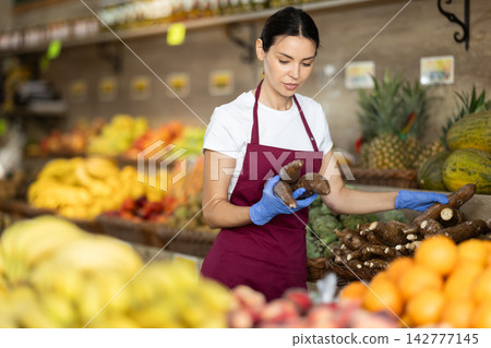 Girl shop seller add cassava goods on display case in vegetable store Girl shop seller add cassava goods on display case in vegetable store 142777145