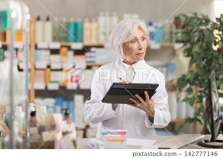 Mature female pharmacist makes notes on electronic tablet - keeps track of receipt and expenditure of pharmacy Mature female pharmacist makes notes on electronic tablet - keeps track of receipt and expenditure of pharmacy 142777146