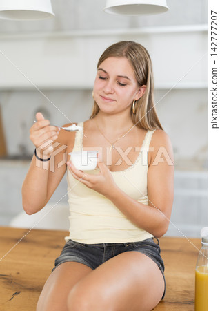 Teenage girl eating fresh yogurt with spoon in modern kitchen Teenage girl eating fresh yogurt with spoon in modern kitchen 142777207