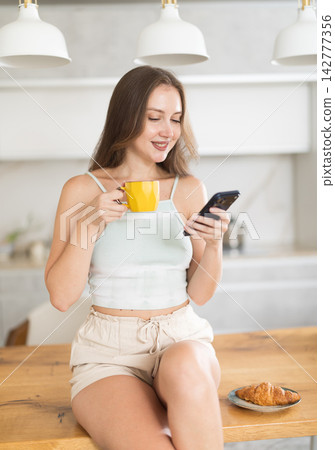 Positive adult woman looking at smartphone while drinking coffee in kitchen Positive adult woman looking at smartphone while drinking coffee in kitchen 142777356