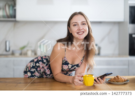 Woman in dress standing in kitchen, looking at mobile phone and drinking coffee 142777373