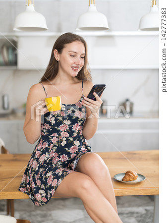 Woman in dress standing in kitchen, looking at mobile phone and drinking coffee Woman in dress standing in kitchen, looking at mobile phone and drinking coffee 142777383