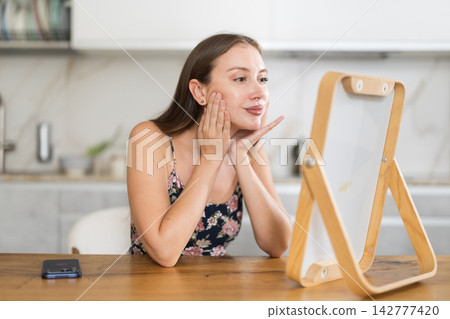 Woman looking at her face in the mirror against the background of the kitchen Woman looking at her face in the mirror against the background of the kitchen 142777420