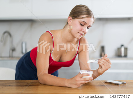 Teenage girl standing in kitchen eating yogurt 142777457