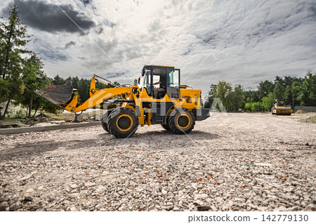 A loader is moving earth and gravel in a rural area to prepare the ground for landscaping. The equipment is active and positioned on a gravel surface A loader is moving earth and gravel in a rural area to prepare the ground for landscaping. The equipment is active and positioned on a gravel surface 142779130