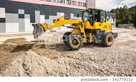 A loader operates at a construction site, moving dirt and gravel to prepare the area for landscaping near a modern building in an urban setting A loader operates at a construction site, moving dirt and gravel to prepare the area for landscaping near a modern building in an urban setting 142779132