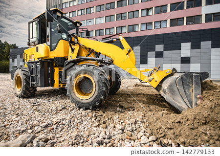 Loader is seen working on a construction site, pushing dirt and gravel. The machine is positioned on rough terrain with buildings in the background Loader is seen working on a construction site, pushing dirt and gravel. The machine is positioned on rough terrain with buildings in the background 142779133