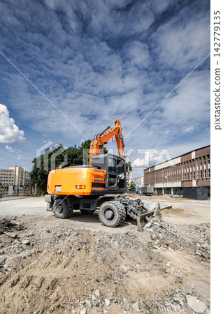 An excavator is digging and moving earth on a landscaping project. The scene shows a construction site with buildings in the background An excavator is digging and moving earth on a landscaping project. The scene shows a construction site with buildings in the background 142779135