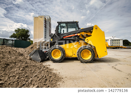 A mini loader operates on a landscaping task, moving earth at a rental location near a tall building. The machine is focused on its work A mini loader operates on a landscaping task, moving earth at a rental location near a tall building. The machine is focused on its work 142779136