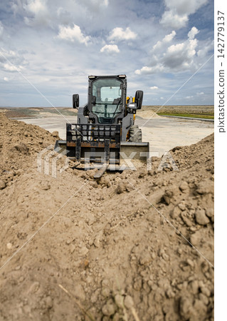 A mini loader is at work on a landscaping project, moving dirt and shaping the terrain at a construction site. The equipment is actively involved in the task A mini loader is at work on a landscaping project, moving dirt and shaping the terrain at a construction site. The equipment is actively involved in the task 142779137