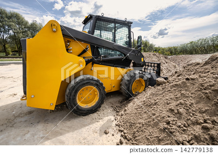 A mini loader is working on a landscaping site, moving earth and gravel to prepare for project completion. It is parked near a pile of dirt A mini loader is working on a landscaping site, moving earth and gravel to prepare for project completion. It is parked near a pile of dirt 142779138