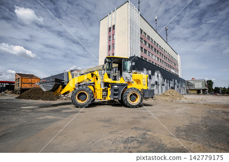 A loader is working at a construction site near a large building, moving dirt and materials for various landscaping tasks A loader is working at a construction site near a large building, moving dirt and materials for various landscaping tasks 142779175