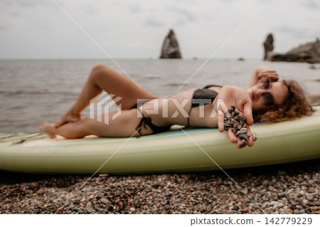Pebbles held by woman lying on paddleboard near rocky ocean shoreline Pebbles held by woman lying on paddleboard near rocky ocean shoreline 142779229