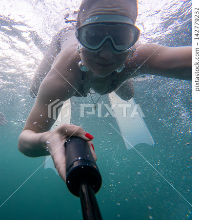 Underwater selfie of woman swimming with goggles and flippers holding action camera in clear sea Underwater selfie of woman swimming with goggles and flippers holding action camera in clear sea 142779232