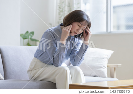 一位四十多歲的婦女坐在沙發上,雙手摀著太陽穴。 一位四十多歲的婦女坐在沙發上,雙手摀著太陽穴。 142780970