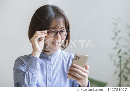 一位四十多歲的女性戴著眼鏡,正在看著她的智慧型手機螢幕。 一位四十多歲的女性戴著眼鏡,正在看著她的智慧型手機螢幕。 142781027